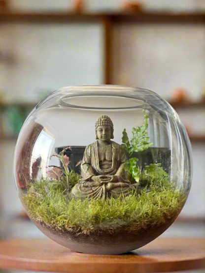 Glass terrarium with a Buddha statue and greenery on a wooden surface.
