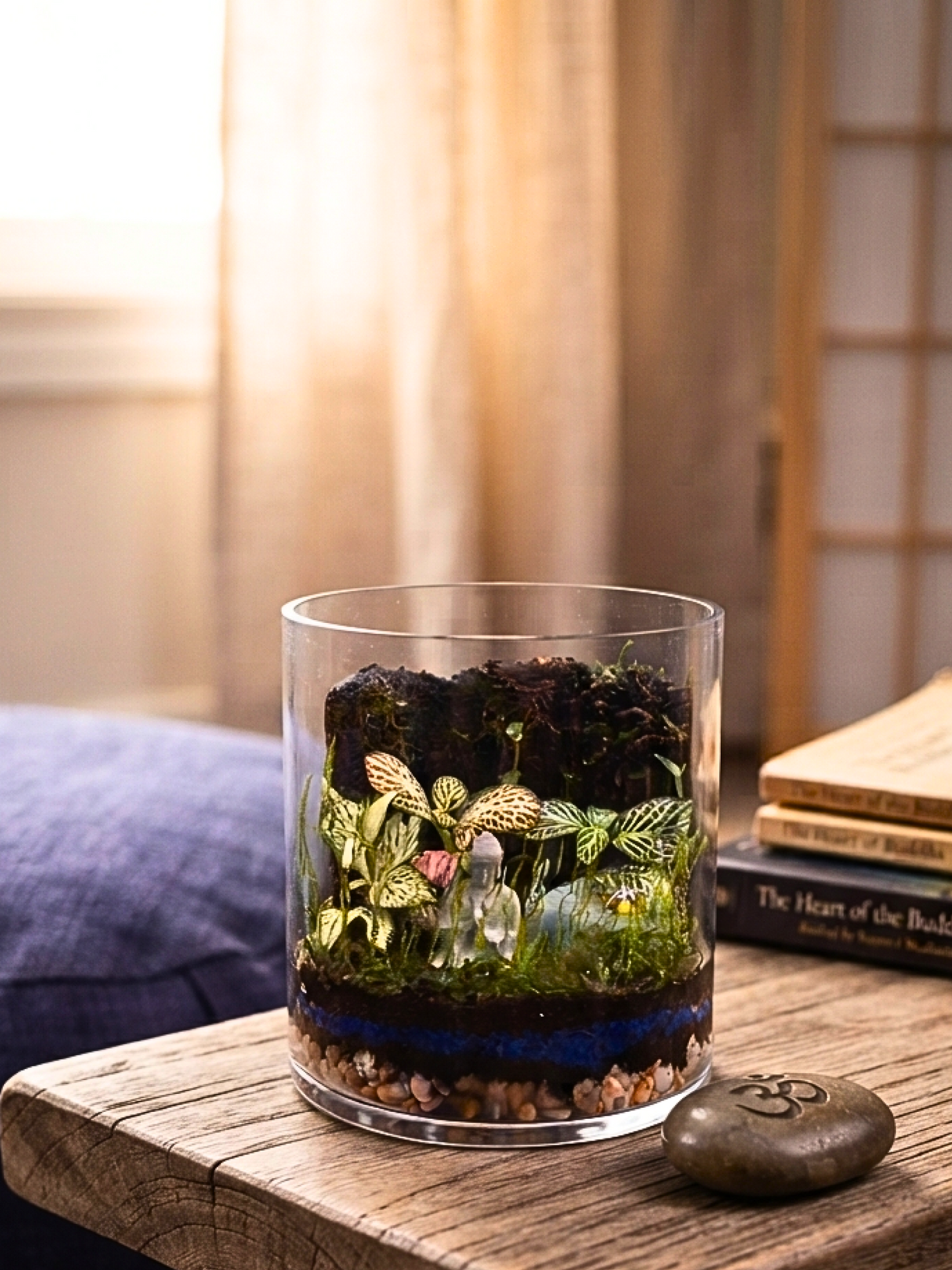 Zen terrarium with Buddha statue, ladybug, blue substrate layer and lush indoor plants in cylinder glass