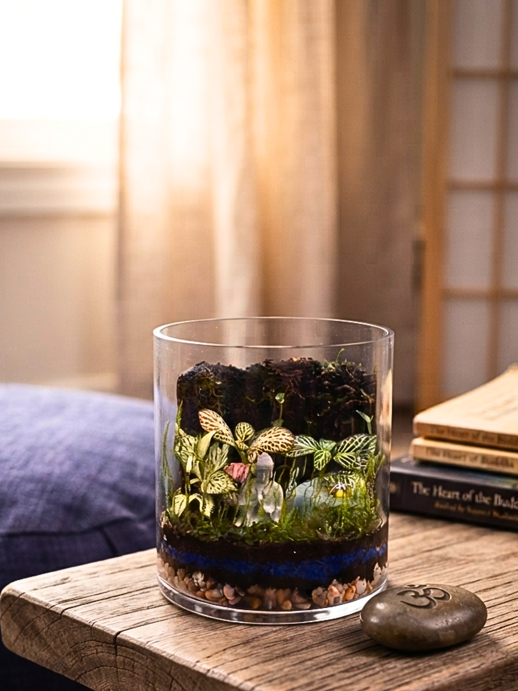 Zen terrarium with Buddha statue, ladybug, blue substrate layer and lush indoor plants in cylinder glass